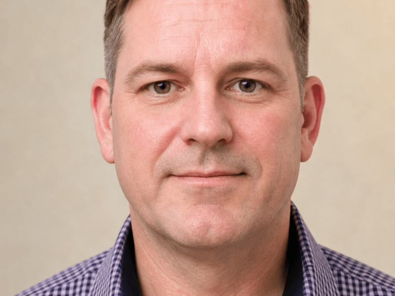 Close-up portrait of a man with short brown hair, wearing a purple checked shirt, against a beige backdrop.