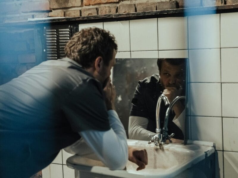Man leaning on a tiled bathroom sink, gazing at his reflection in the mirror with a thoughtful expression.