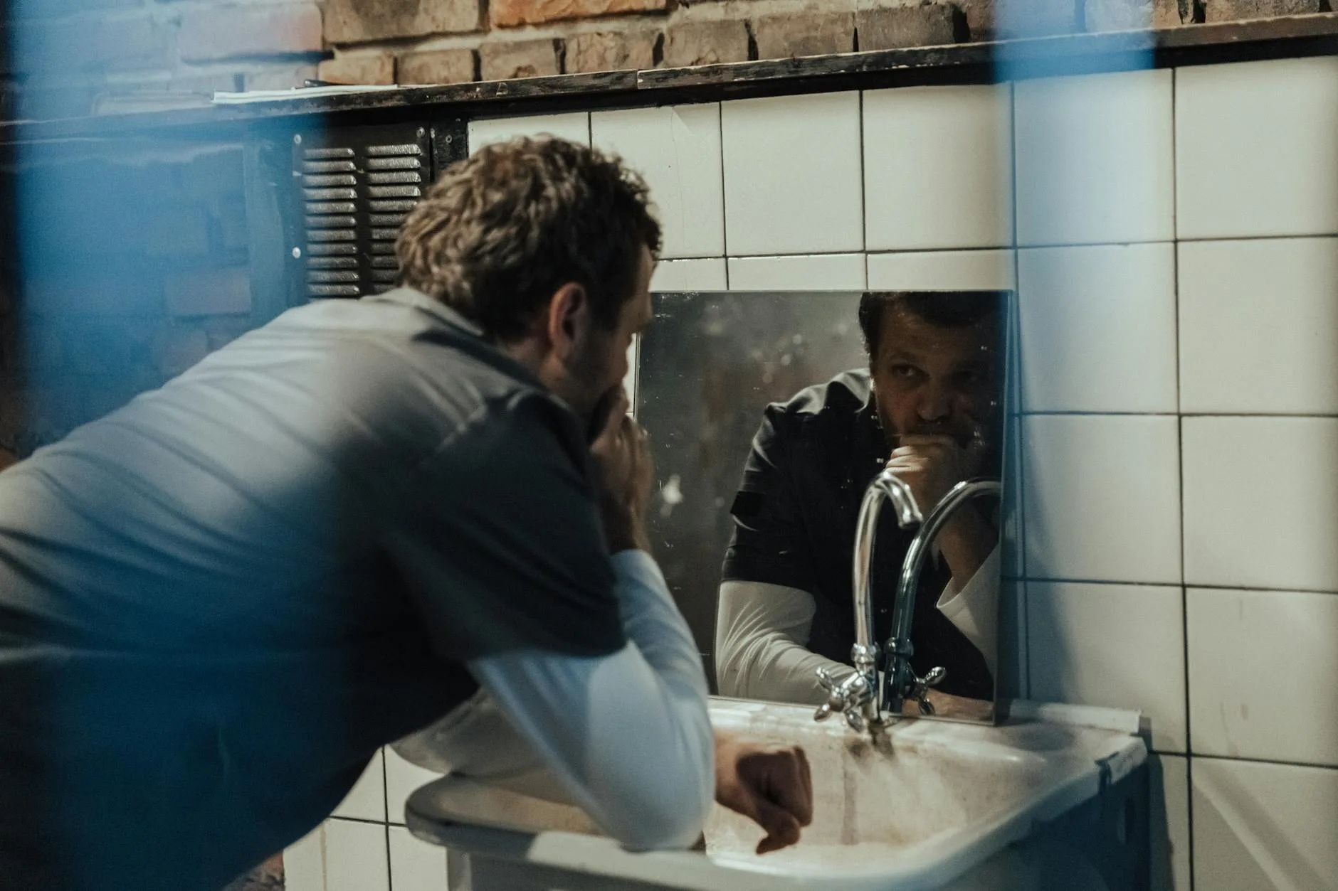 Adult man leaning over a tiled bathroom sink and looking thoughtfully into a mirror