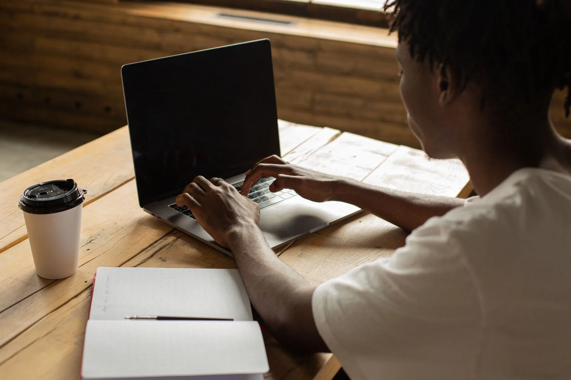 A person at a laptop with an open notebook and takeaway cup on the table.