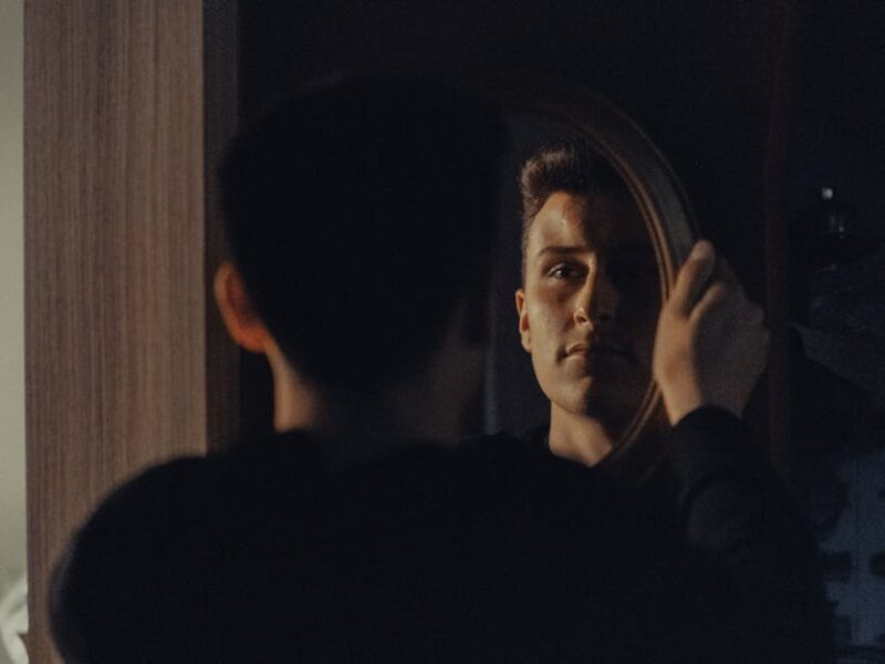 Young man in a dim room looks at his reflection in an oval mirror, holding the frame with one hand.