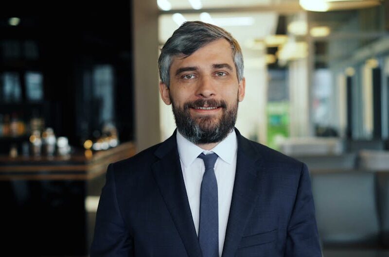 Smiling man in a navy suit and tie stands in a modern office lobby or lounge area.