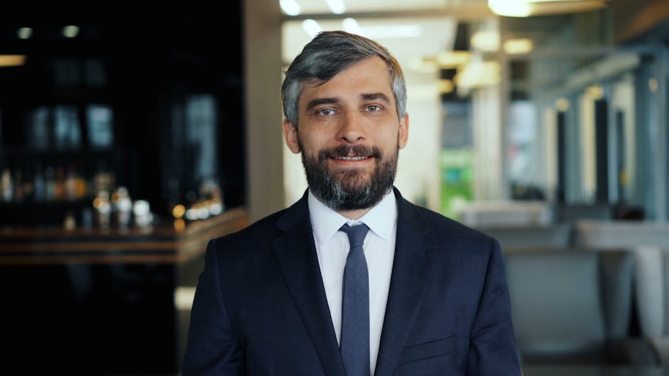 Smiling man in a navy suit and tie stands in a modern office lobby or lounge area.