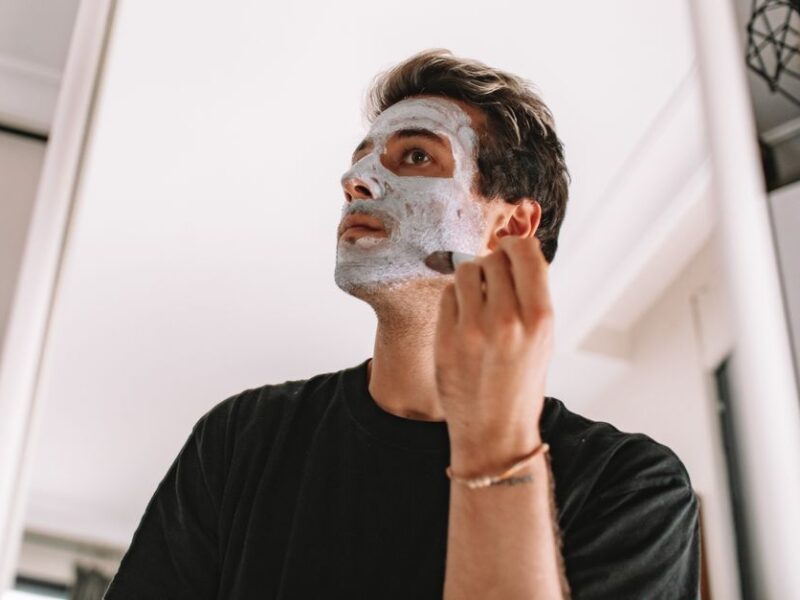 Man applying a white facial mask at home, looking upward in a bright bathroom setting.