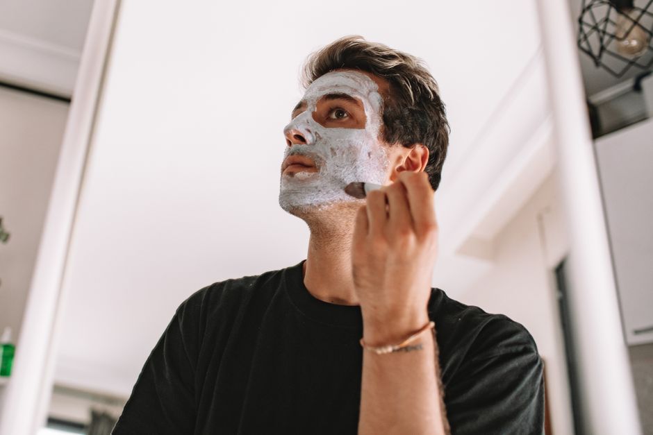 Man applying a white facial mask at home, looking upward in a bright bathroom setting.