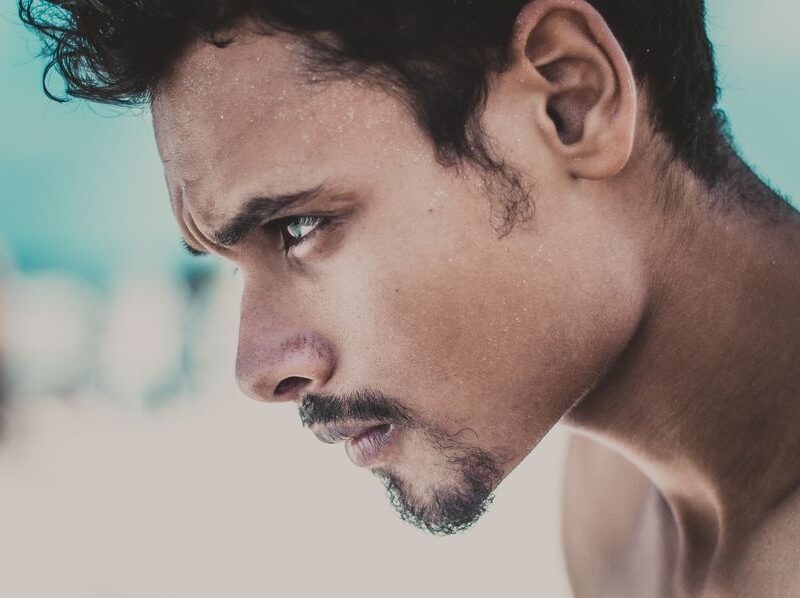 Close-up profile of a man with short facial hair looking downward on a sunny beach background.
