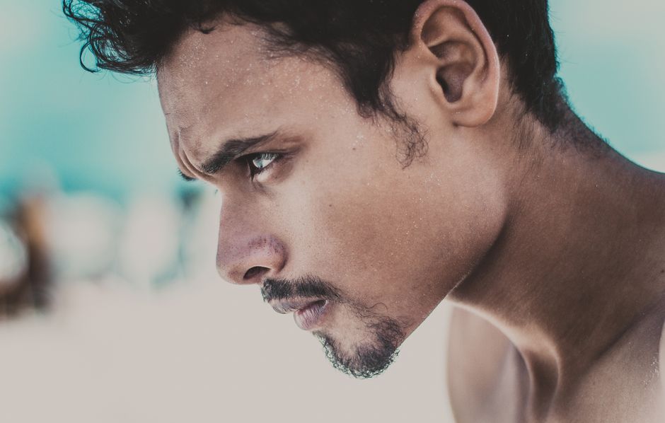 Close-up profile of a man with short facial hair looking downward on a sunny beach background.