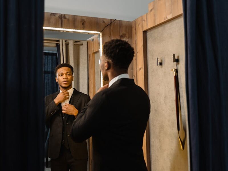 Man in a suit adjusting his tie while looking at himself in a mirror in a dressing room with wooden walls and blue curtains