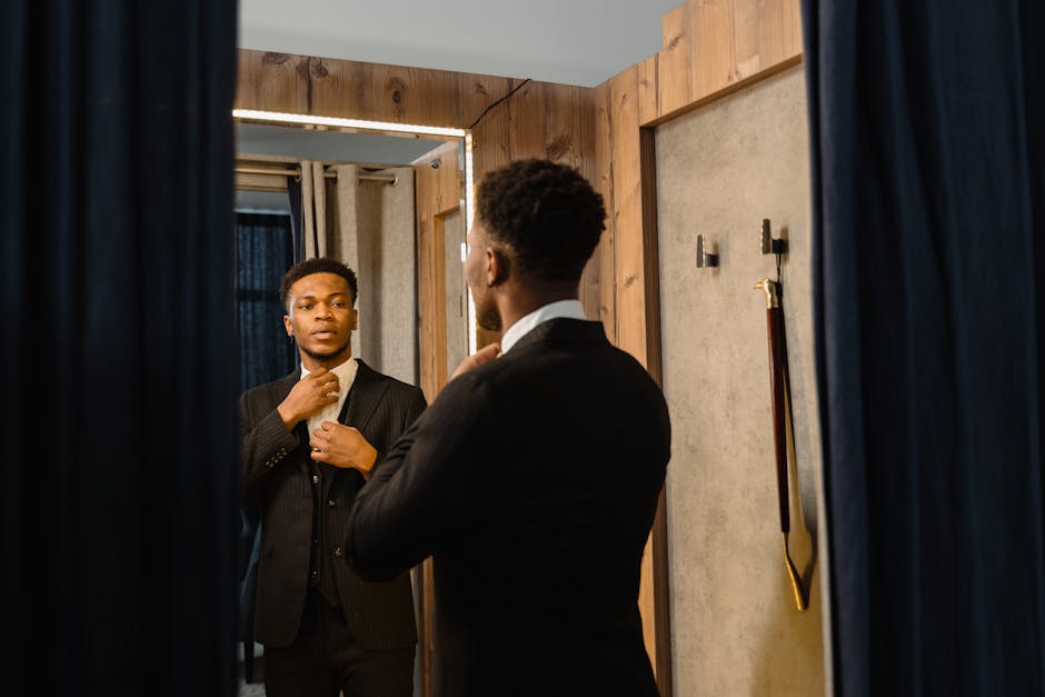 Man in a suit adjusting his tie while looking at himself in a mirror in a dressing room with wooden walls and blue curtains