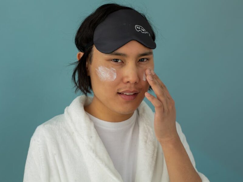 Person wearing a sleep mask and white robe applies facial cream to their cheeks against a teal background.