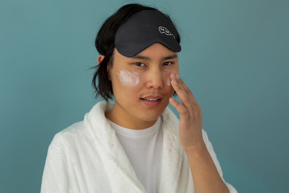 Person wearing a sleep mask and white robe applies facial cream to their cheeks against a teal background.