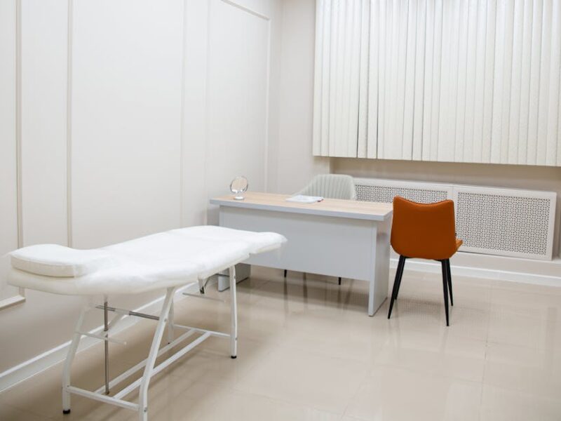 Examination room with a white medical couch, a light desk, orange chair, and vertical blinds.
