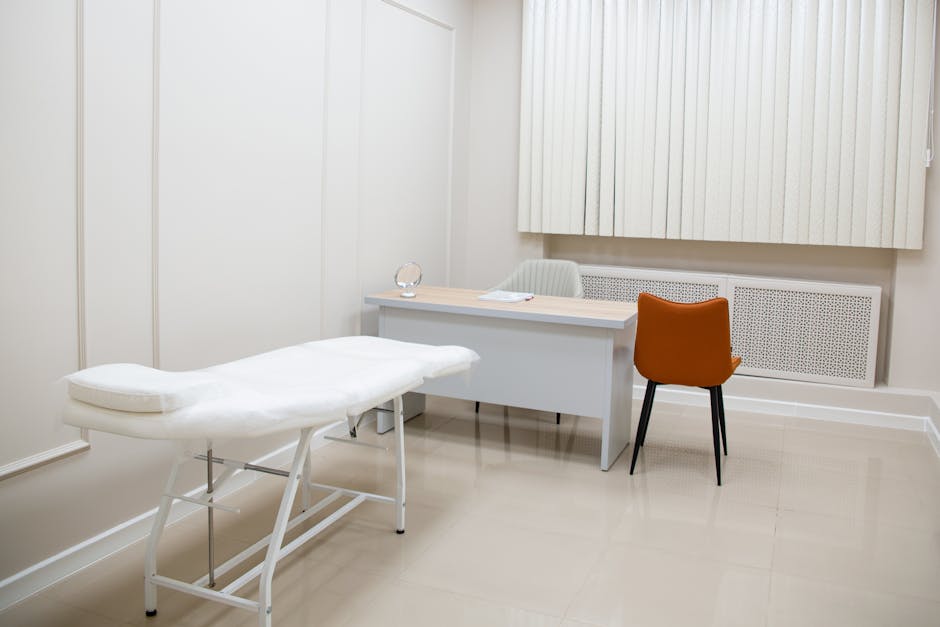 Examination room with a white medical couch, a light desk, orange chair, and vertical blinds.