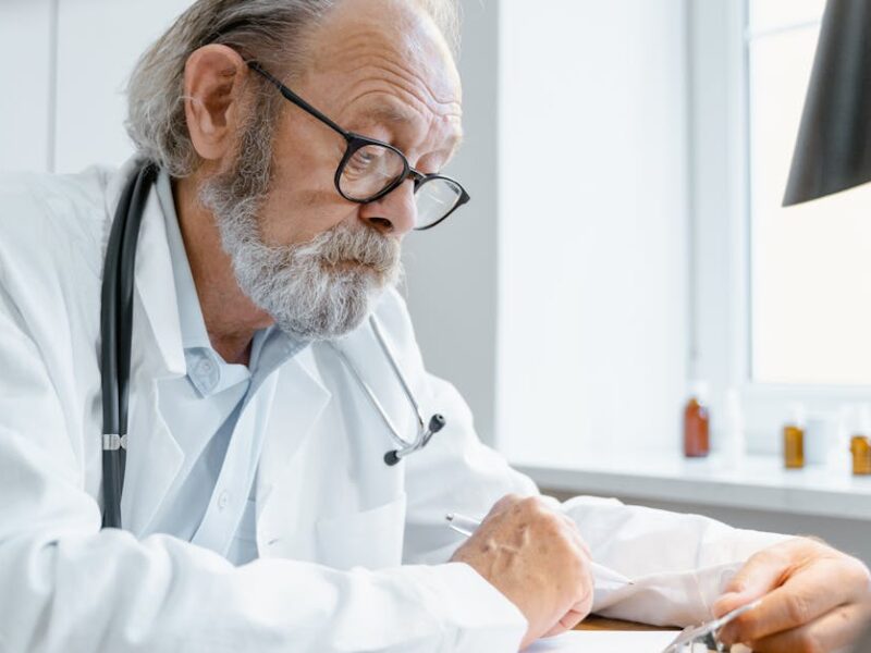 Elderly male doctor with glasses and stethoscope writing at a bright desk in a clinic.