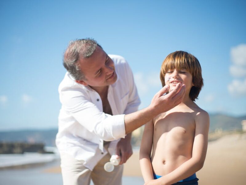 Adult man in a white shirt at the beach feeds a snack to a shirtless boy as they smile under a sunny sky by the ocean.