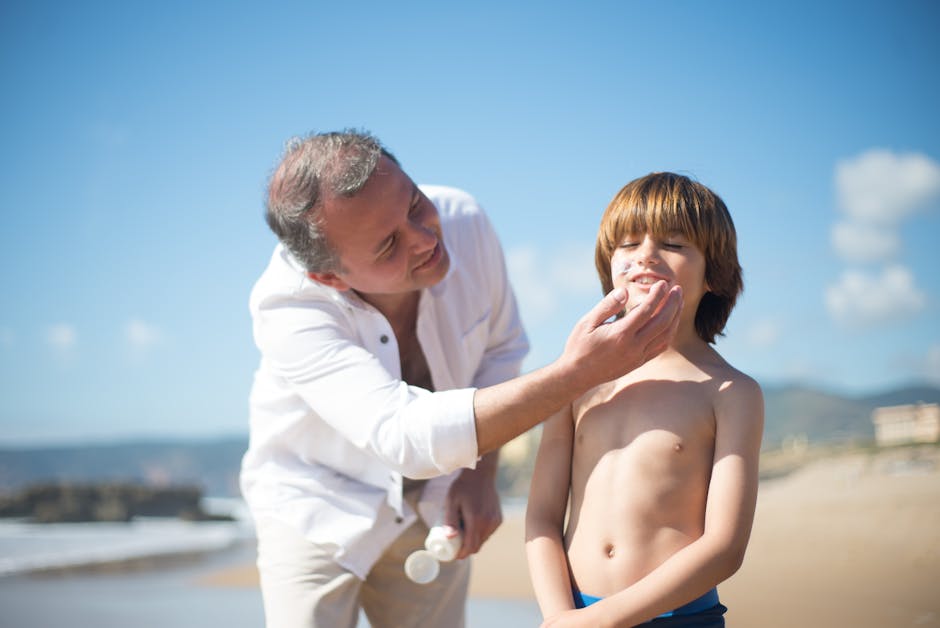 Adult man in a white shirt at the beach feeds a snack to a shirtless boy as they smile under a sunny sky by the ocean.